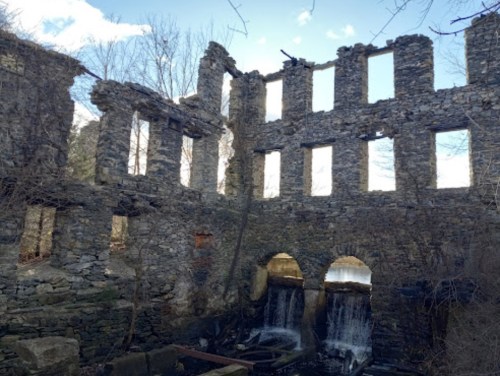 Ruins of a stone building with arched openings, surrounded by trees and a small waterfall in the foreground.