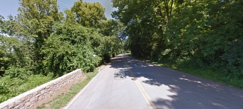 A quiet, tree-lined road with a stone wall on one side, surrounded by lush greenery.