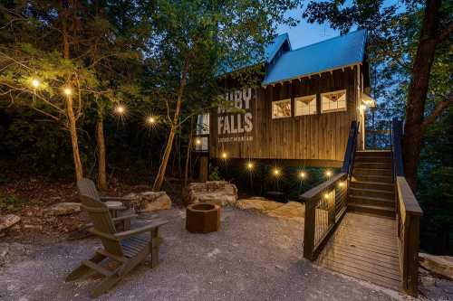 A cozy cabin with a sign reading "Cypress Falls," surrounded by trees and lit by string lights, with a fire pit nearby.