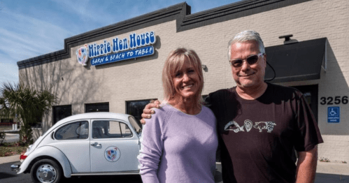 A man and woman smile in front of a restaurant called "Hippie Hen House," with a vintage car parked nearby.