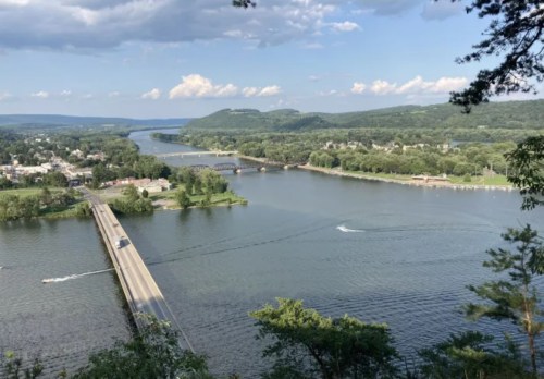 A scenic view of a river winding through green hills, with a bridge and small town visible along the shoreline.
