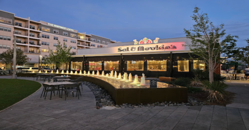 A restaurant with a fountain in front, surrounded by greenery, under a twilight sky. Modern buildings in the background.