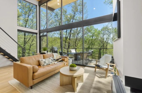 Modern living room with a tan leather sofa, round coffee table, and large windows overlooking a green outdoor view.