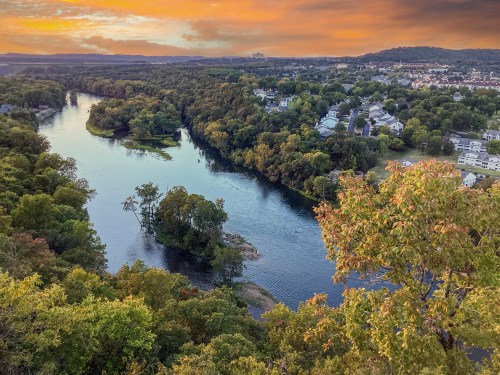 A scenic view of a river winding through lush greenery and homes, with a vibrant sunset sky in the background.