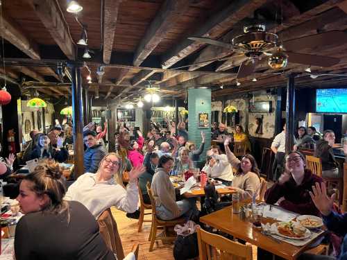 A lively restaurant scene with many people smiling and waving, seated at tables with food and drinks.