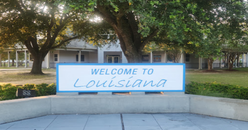 Sign reading "Welcome to Louisiana" in front of trees and a building.