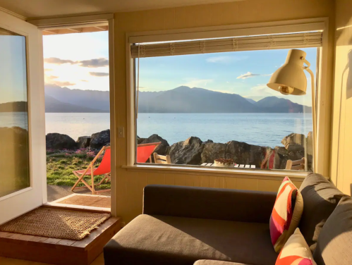 Cozy interior with a sofa, lamp, and a stunning view of mountains and water through a large window.