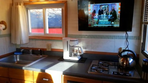 A cozy kitchen with a sink, coffee maker, kettle, and a TV showing a talk show on the wall. Natural light streams in.