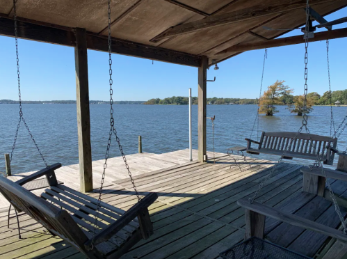 A wooden dock with swings overlooking a calm lake on a clear day. Trees are visible in the distance.