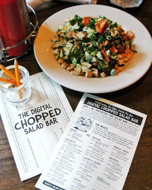 A colorful chopped salad on a plate next to a menu for a digital salad bar, featuring various ingredients and rules.