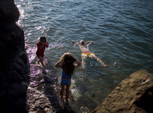 Three children play by the water; one floats while the others stand on the rocks, enjoying a sunny day.