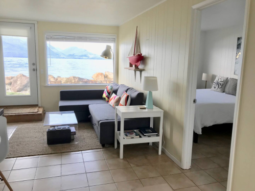 Cozy interior with a living area, a bedroom visible, and a view of the water through large windows.