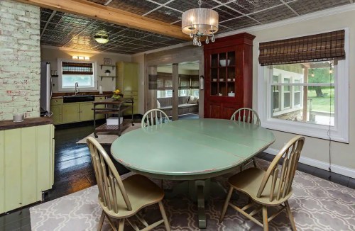 A cozy dining area with a round green table, four chairs, and a chandelier, featuring a view of a sunny kitchen.