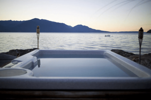 A serene hot tub overlooking a calm lake at sunset, with mountains in the background and tiki torches nearby.
