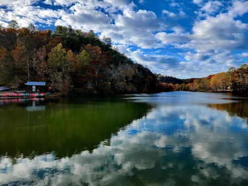 A serene lake surrounded by autumn trees, reflecting clouds and vibrant colors under a blue sky.