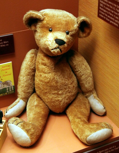 A vintage teddy bear with a brown fur coat, sitting upright against a wooden background.