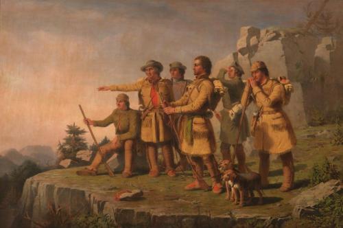 A group of six explorers in fur coats stands on a rocky outcrop, gazing into the distance with two dogs nearby.