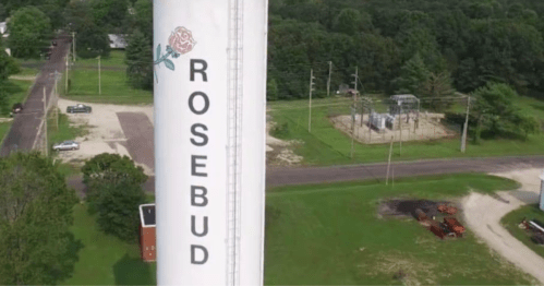 Aerial view of a water tower with "ROSEBUD" and a rose graphic, surrounded by greenery and roads.