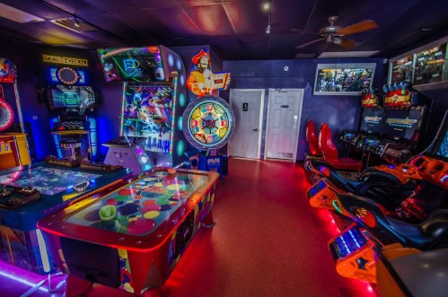A vibrant arcade room filled with colorful games, including air hockey, racing, and a prize wheel, illuminated by neon lights.