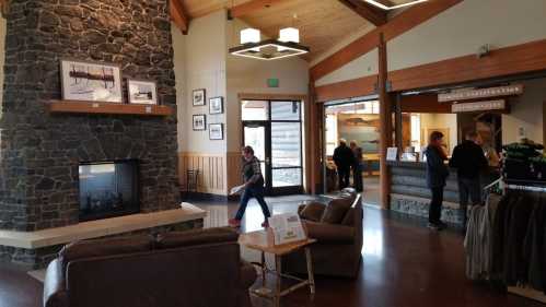 A cozy lodge interior with a stone fireplace, wooden beams, and visitors at the reception area.