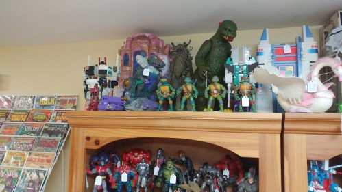A shelf displaying various action figures, including dinosaurs and superheroes, alongside comic books in the background.
