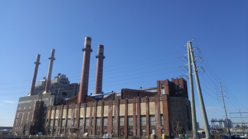 A large industrial building with three smokestacks against a clear blue sky, surrounded by power lines.