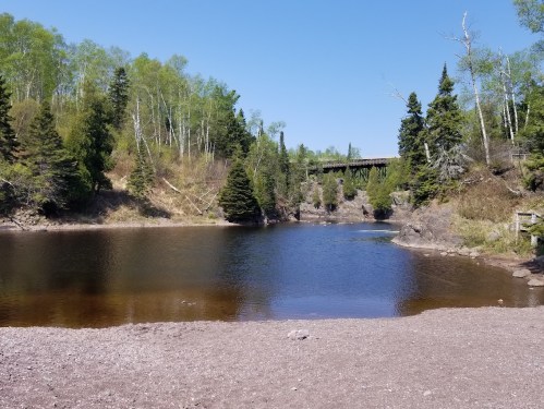 A serene landscape featuring a calm river, lush green trees, and a bridge in the background under a clear blue sky.