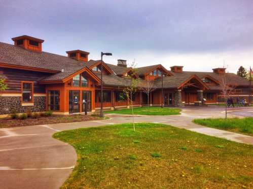 A modern wooden building with a sloped roof, surrounded by green grass and a paved pathway under a cloudy sky.