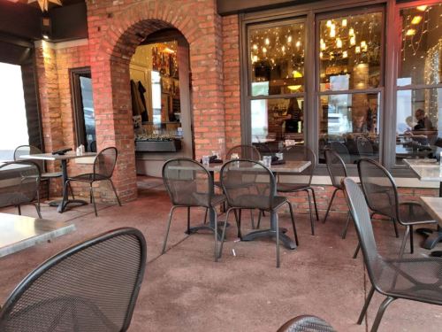 Outdoor dining area with empty tables and chairs, featuring brick arches and warm lighting in the background.