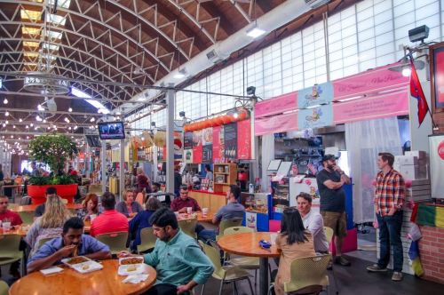 A bustling food hall with people dining at tables, colorful stalls, and vibrant decor in a lively atmosphere.