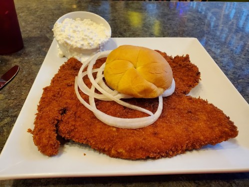 A large fried fish fillet with sliced onions, a dinner roll, and a side of cottage cheese on a white plate.