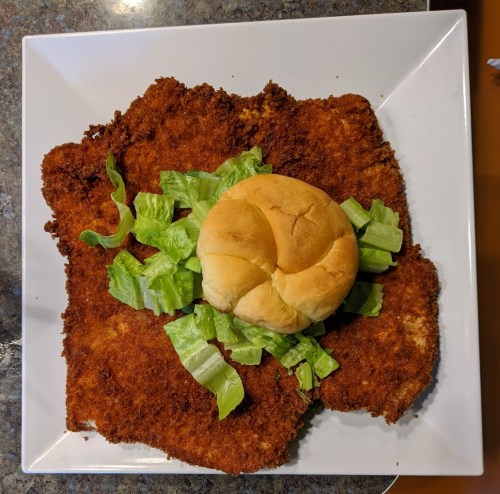 A large, crispy fried chicken cutlet on a plate, topped with lettuce and a soft bun in the center.