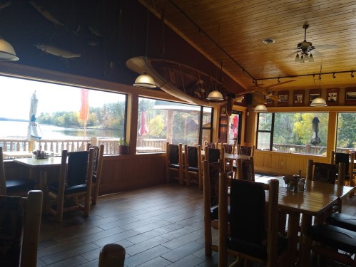 Cozy restaurant interior with wooden decor, large windows overlooking a lake, and empty tables ready for diners.