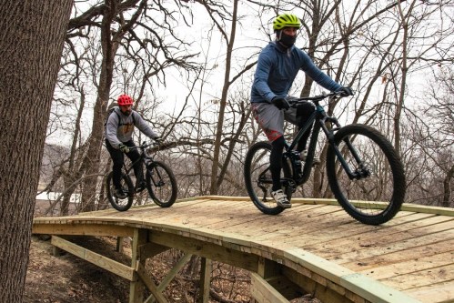 Two mountain bikers ride on a wooden trail through a wooded area, wearing helmets and winter gear.