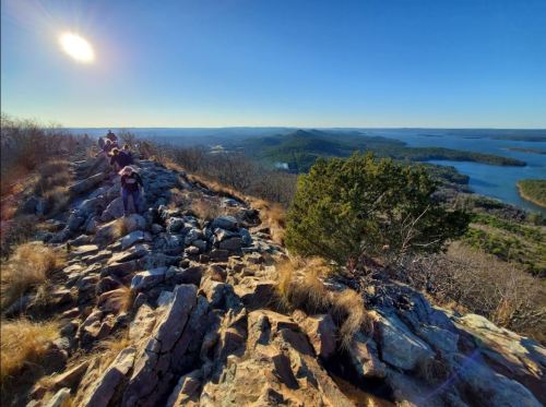 Hikers on a rocky mountain trail with a scenic view of lakes and hills under a clear blue sky.