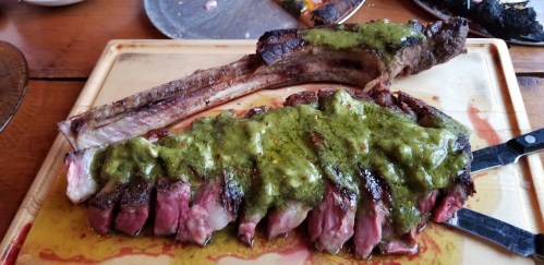 A large, grilled rib steak topped with green sauce, served on a wooden cutting board.
