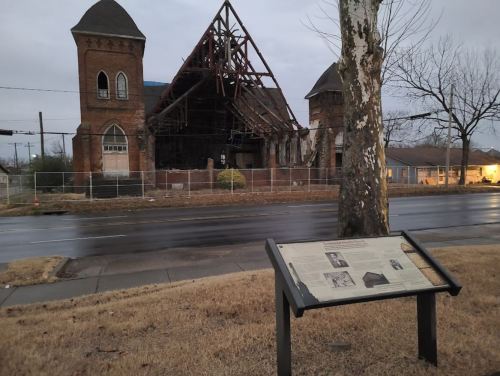A historic church in disrepair, with a nearby informational sign, set against a cloudy sky and empty street.