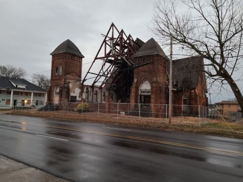 A dilapidated church with a collapsed roof, surrounded by a fence, on a rainy street.
