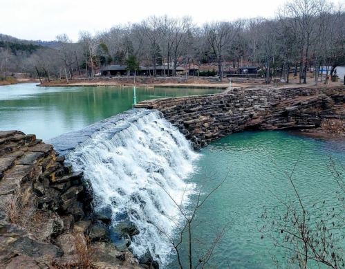 A serene waterfall cascades over a stone structure into a calm, greenish-blue lake surrounded by bare trees.