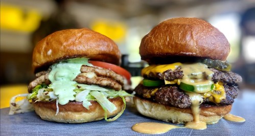 Two gourmet burgers on a gray surface: one with chicken, lettuce, and sauce; the other with beef, cheese, and jalapeños.