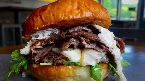 A close-up of a juicy burger with roast beef, melted cheese, lettuce, and creamy sauce on a toasted bun.