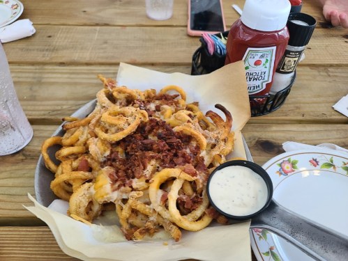 A plate of loaded curly fries topped with cheese and bacon, served with a side of ranch dressing and ketchup.