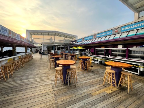 Outdoor dining area with wooden tables and stools, featuring a bar and a sunset in the background.
