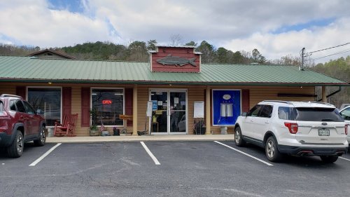 A rustic building with a green roof, featuring a fish sign, open sign, and parked cars in front.