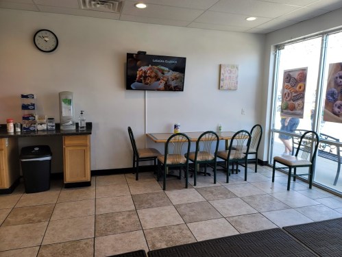 A clean, bright cafe interior with a table, chairs, a clock, and a TV displaying food, plus a view of the outside.