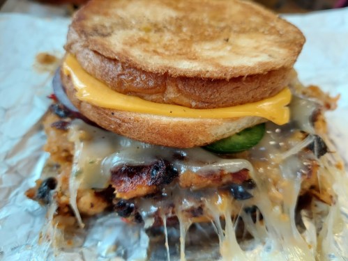 A close-up of a sandwich with grilled chicken, melted cheese, jalapeños, and toasted bread, oozing with toppings.