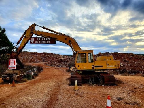 A yellow excavator at a dirt road entrance, with a sign reading "No Vehicles" and a warning sign nearby.