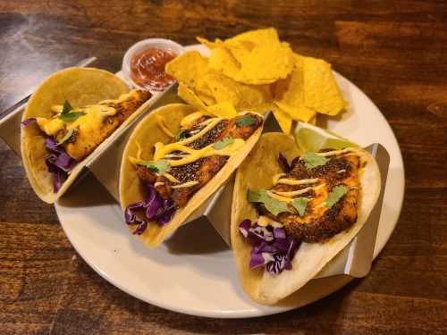 Three tacos with grilled chicken, purple cabbage, and sauce, served with a side of tortilla chips and salsa.