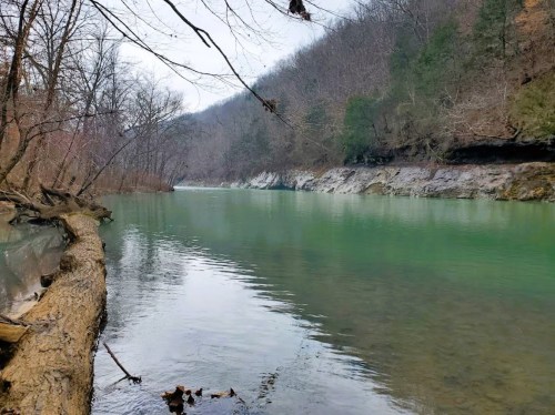 A serene river flows through a wooded landscape, with rocky banks and bare trees lining the water's edge.