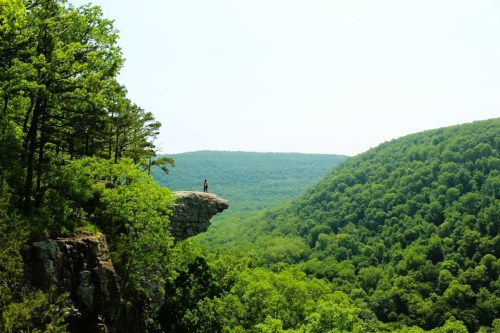 A person stands on a rocky outcrop overlooking a lush green valley and rolling hills under a clear sky.